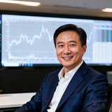 一位穿着深蓝色西装的金融行业技术高管在办公室中微笑的半身肖像照，背景为数据大屏显示实时交易监控曲线
